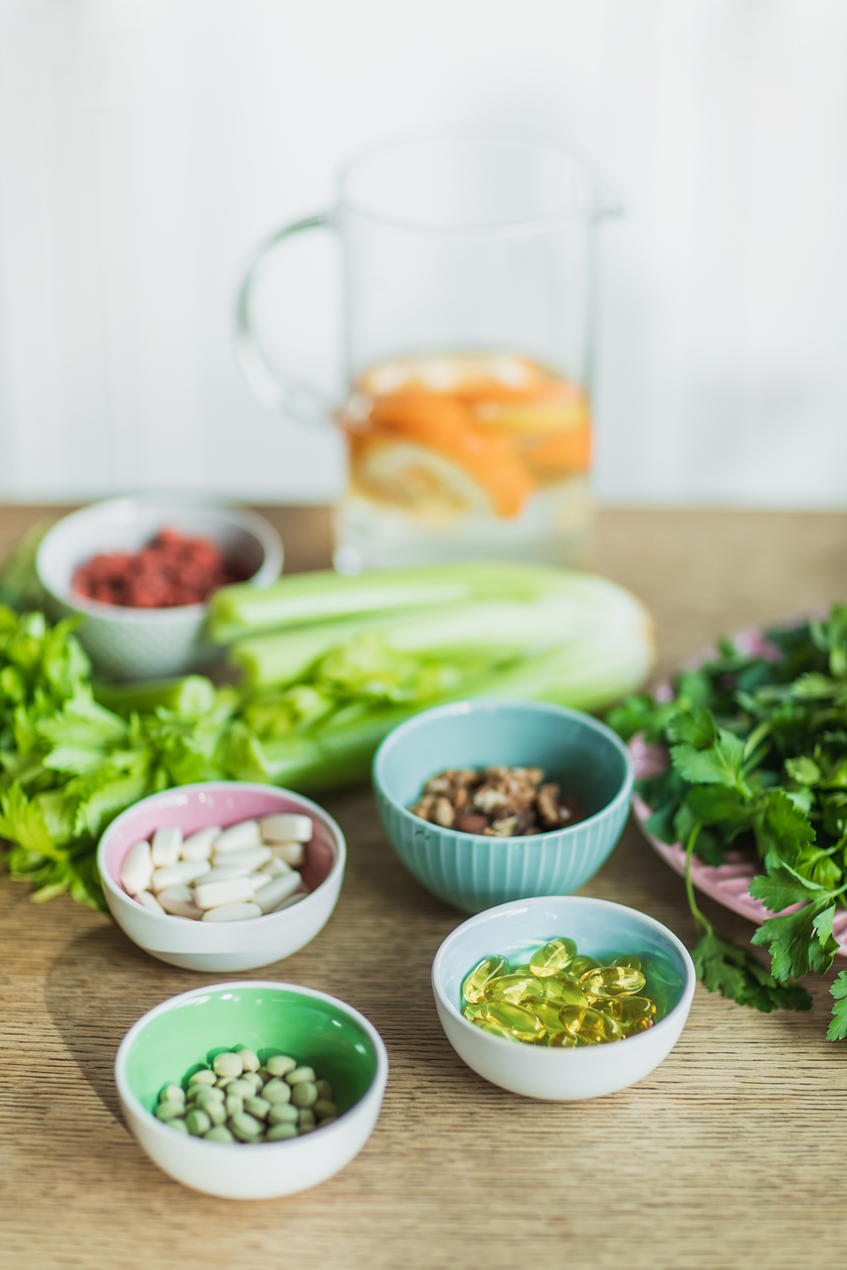 Several small bowls containing vitamins sit on a wooden table with celery and a jug of water in the background.