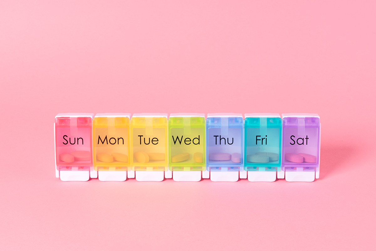 A multi-coloured weekly pill organiser is set against a pink background.