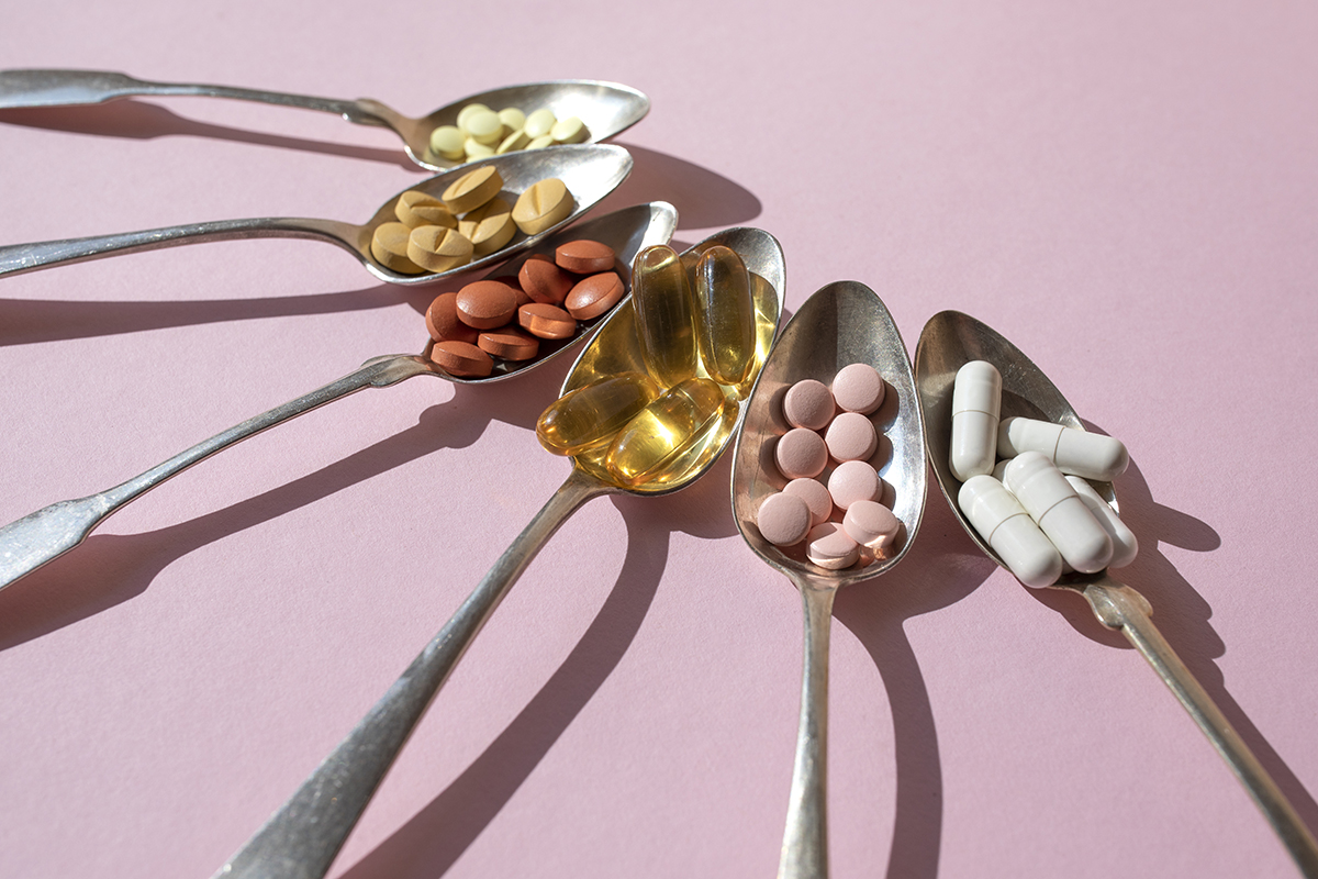 Six metal spoons containing different coloured vitamins are positioned on a pink background.
