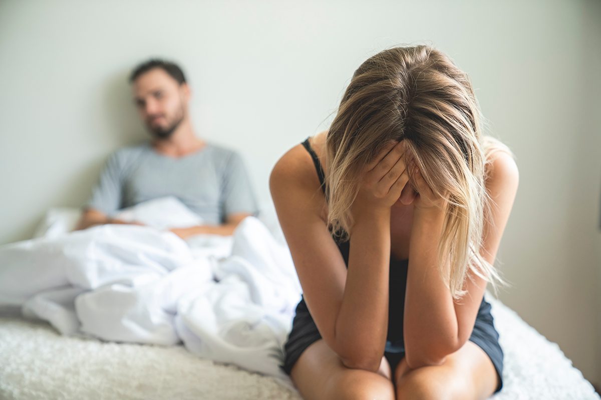 A woman sits on the edge of a bed while her partner sits behind her, both looking away from each other, suggesting conflict