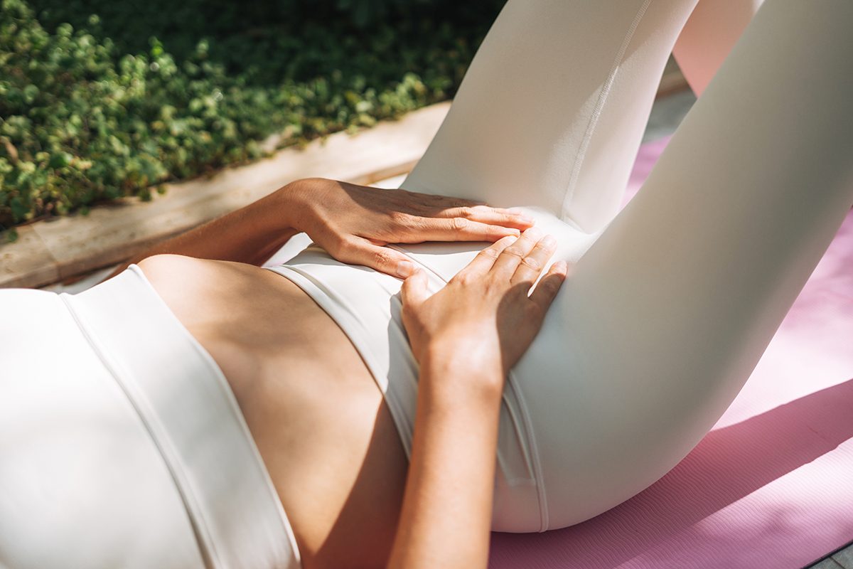 A woman is lying on a yoga mat outdoors, with her hands resting on her lower abdomen