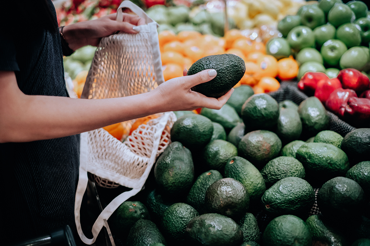 A woman holds an avocado in her hand in the fruit and vegetable aisle of a supermarket.