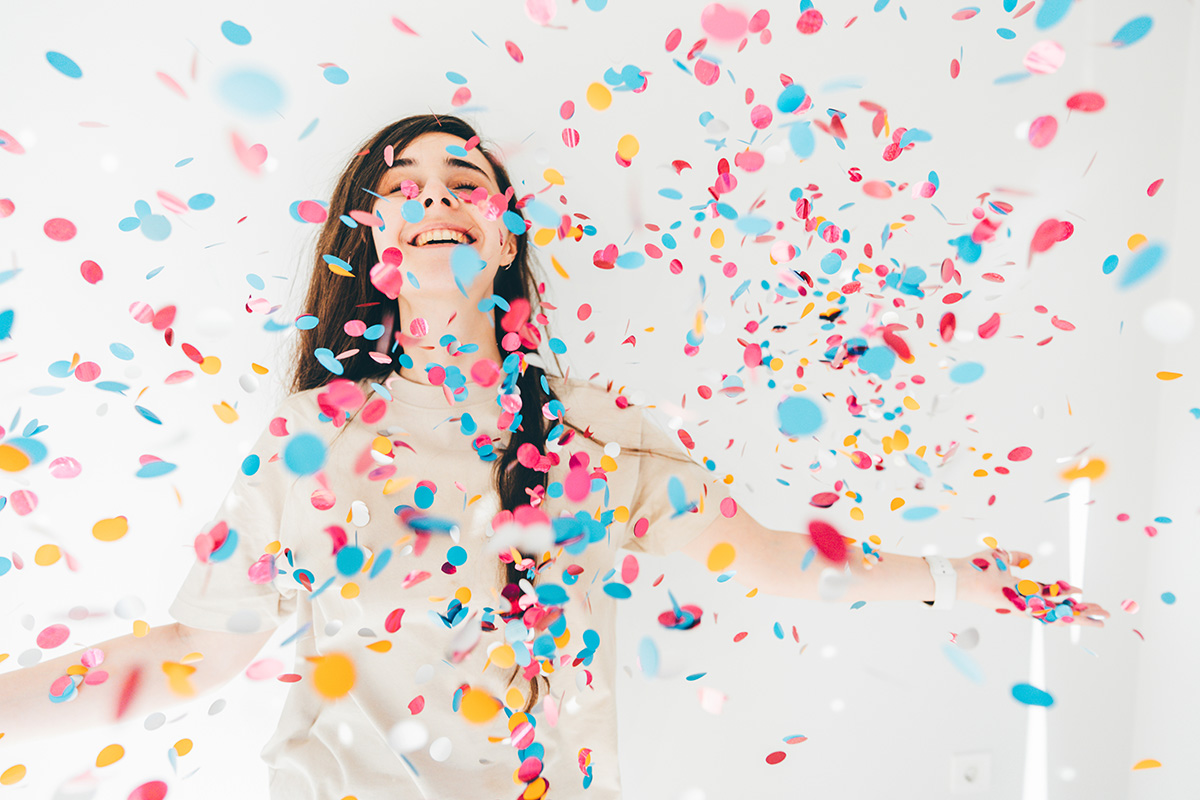 A smiling woman is standing amongst confetti with her arms outstretched