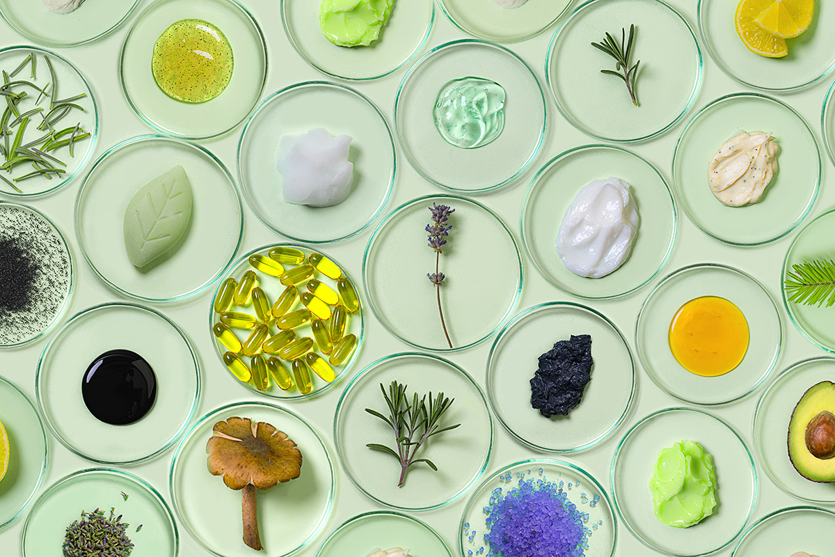 A selection of ingredients, creams, and vitamins are arranged on petri dishes on a green background.