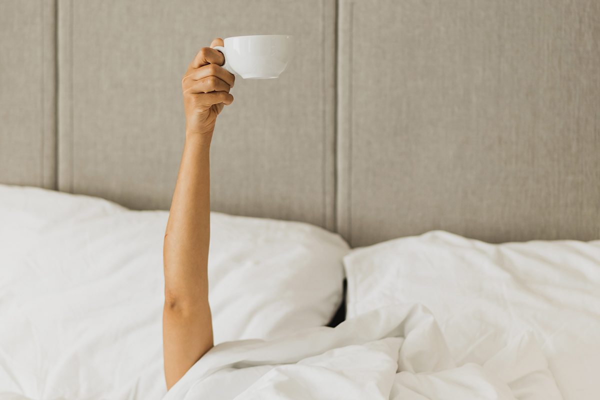 An arm stretches out of a bed with white bedding and holds up a white mug.