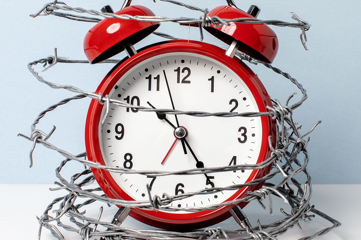 A red alarm clock is wrapped in barbed wire against a light blue background.