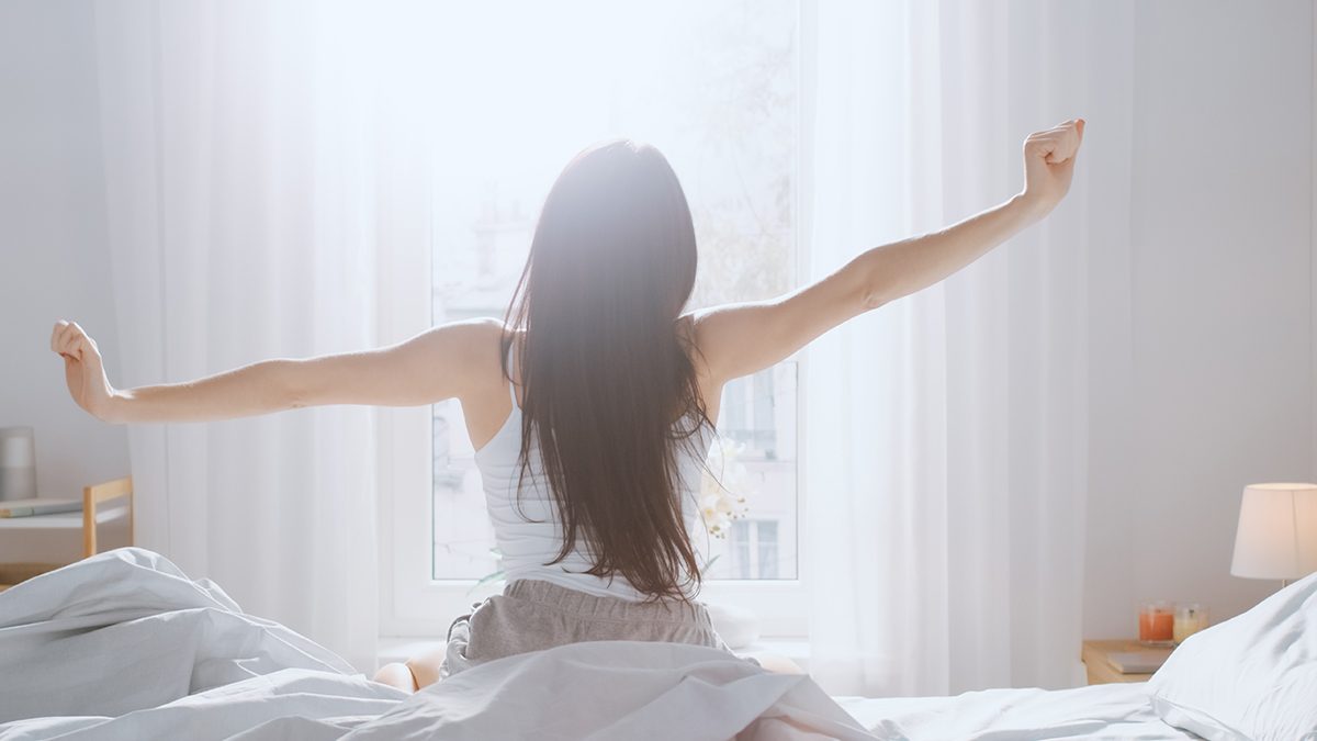 A woman with her back to the camera stretches her arms in bed as sunlight streams through a large window