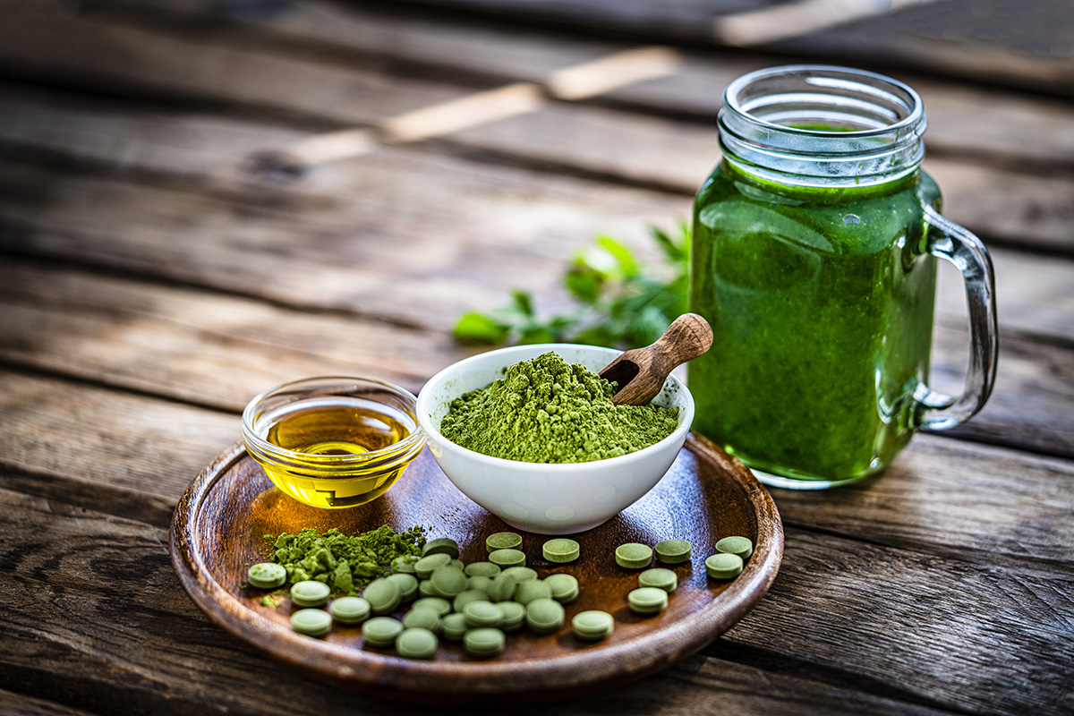 A jar containing a green drink and a round wooden tray holding a small bowl of green powder with a wooden scoop, a dish of green tablets, and a small bowl of oil are set on a wooden surface.
