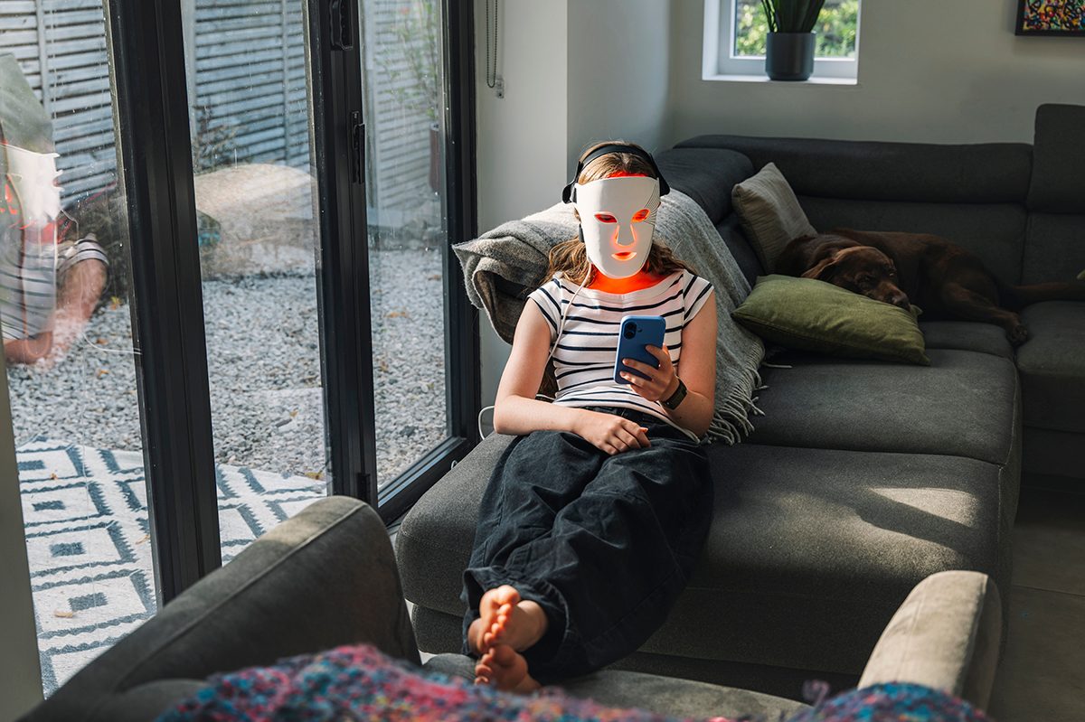 A young girl is sitting on a sofa and looking at her mobile phone while wearing a red light therapy mask