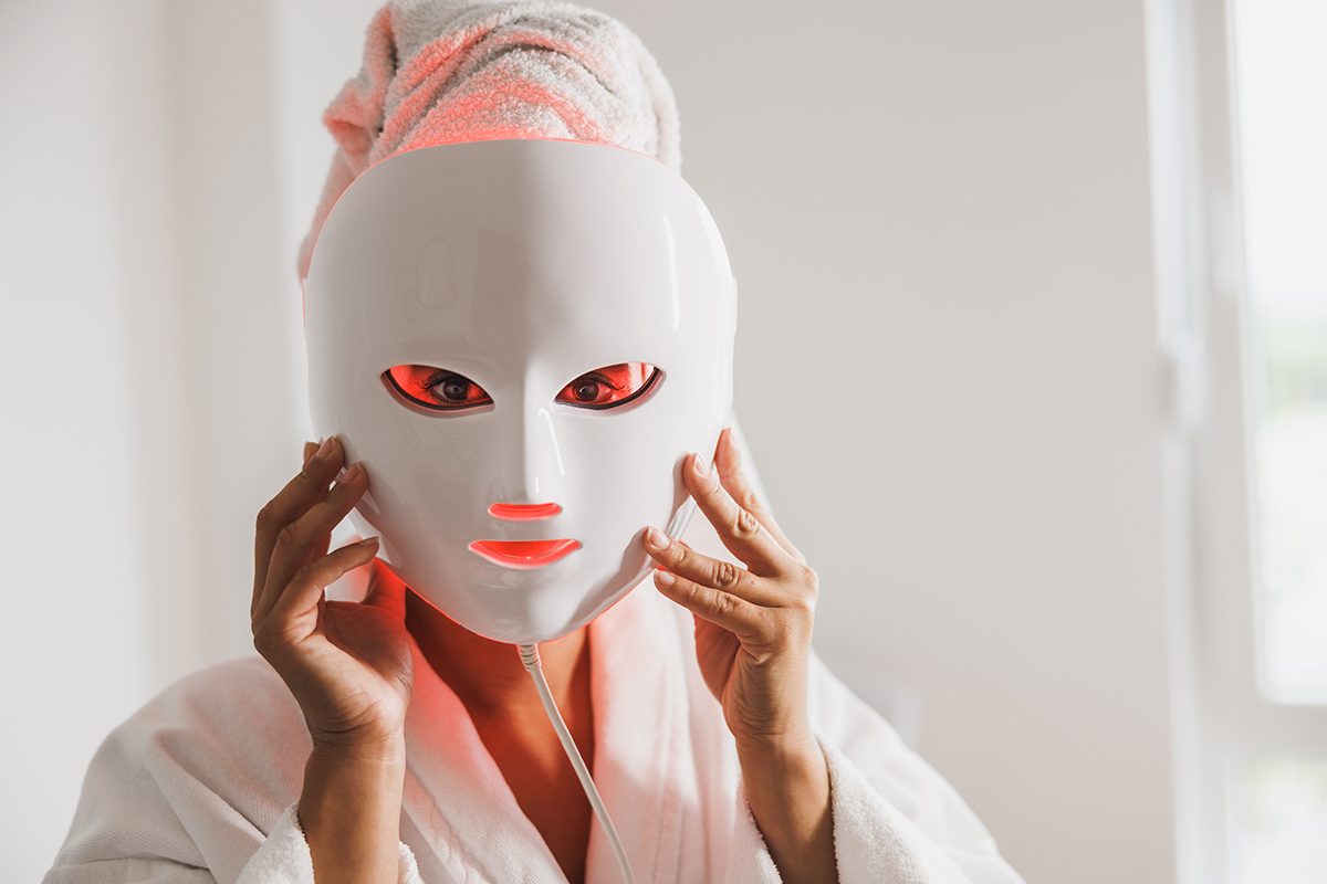 A woman, who has her hair wrapped in a towel and is wearing a bath robe, holds an LED red light therapy mask to her face