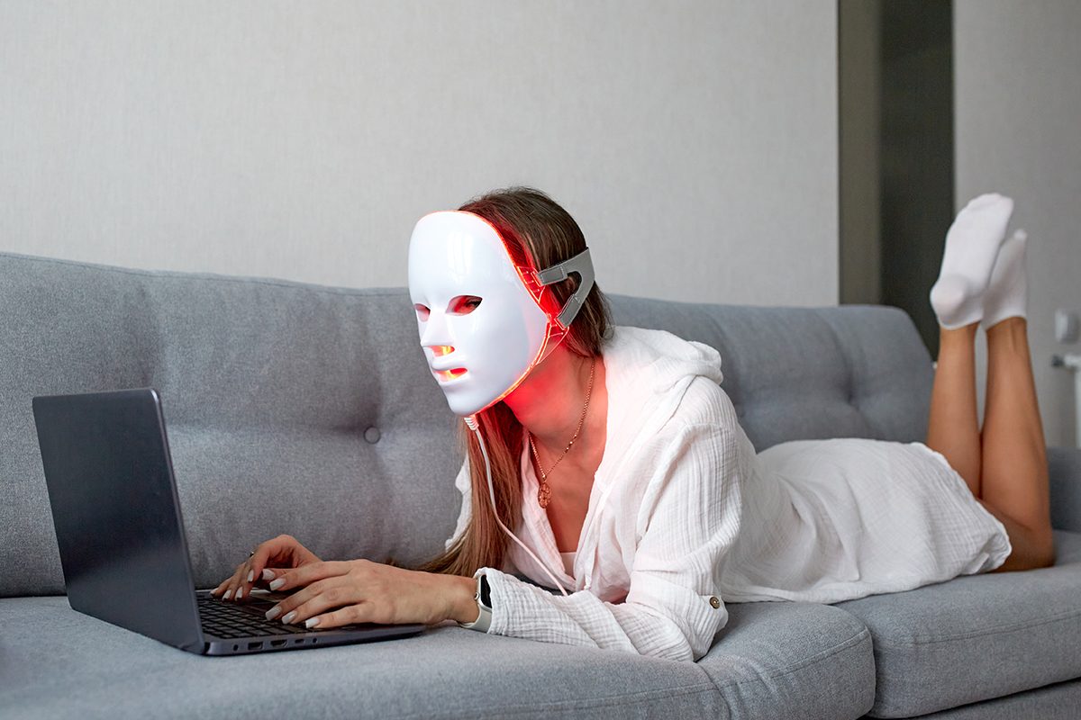 A woman wearing an LED face mask is lying on a sofa and working on a laptop