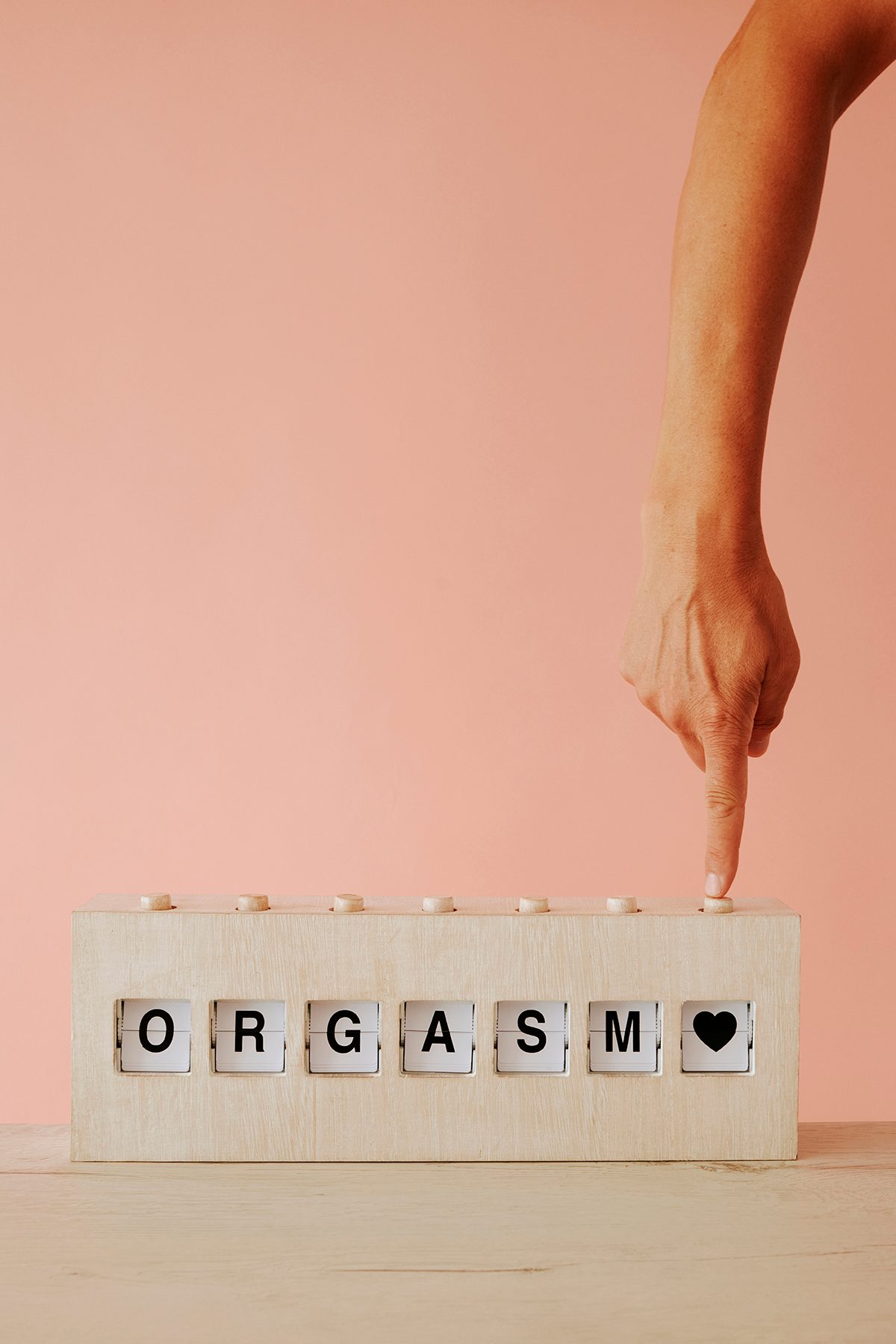 A hand pushes a button on a wooden split-flap display that reads 'ORGASM'