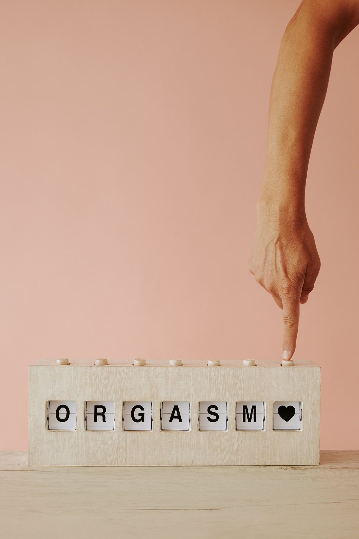 A hand pushes a button on a wooden split-flap display that reads 'ORGASM'