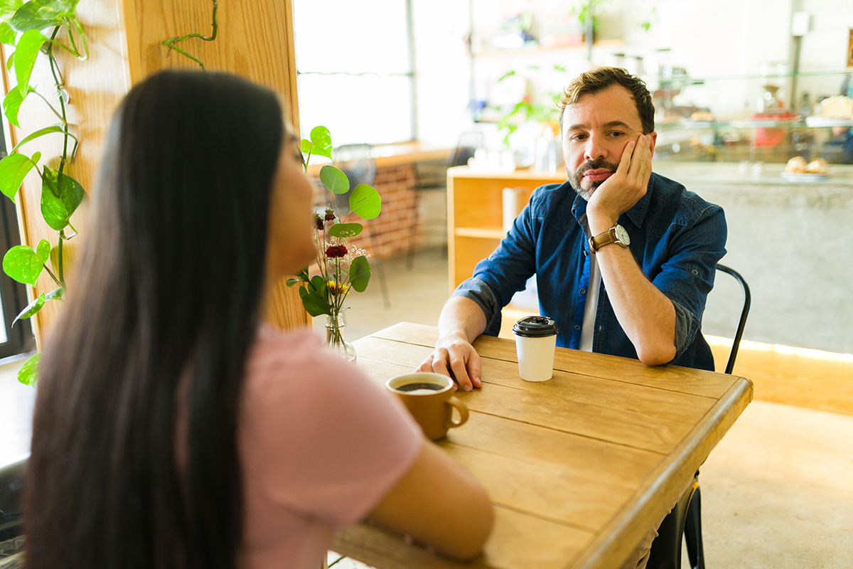 A man sits across a table from a woman in a cafe, looking bored