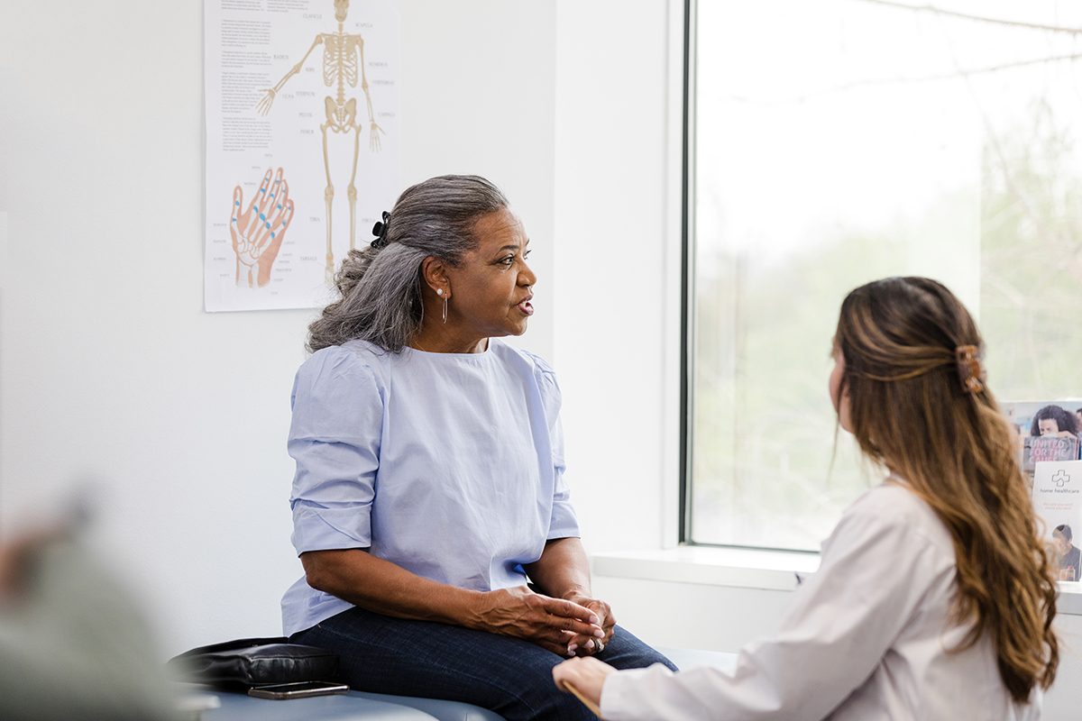 A woman sitting on an examination table talks to a female doctor 