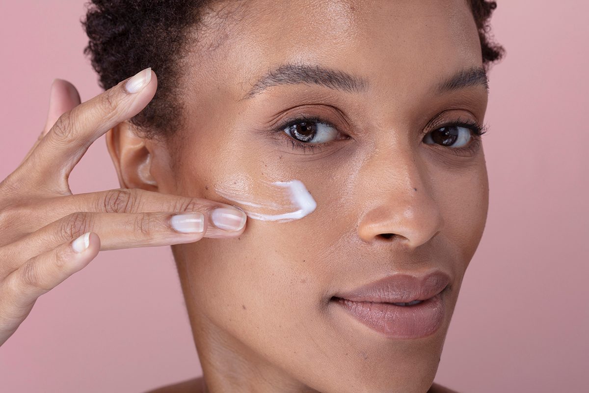 A close-up of a woman applying moisturiser to her face against a pink background