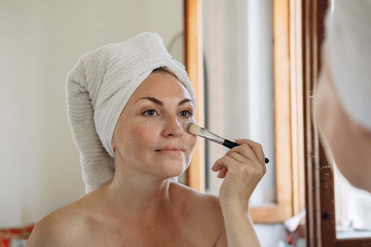 A woman with her hair wrapped in a towel applies foundation to her under-eye area with a brush in the mirror