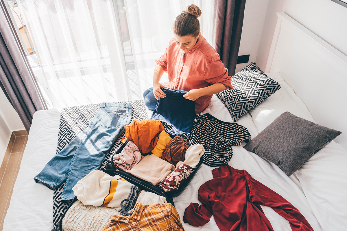 A woman is sitting on the edge of a bed packing a suitcase