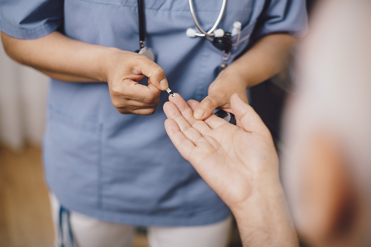 A healthcare professional in blue scrubs is holding a patient's hand to take a blood sugar reading.