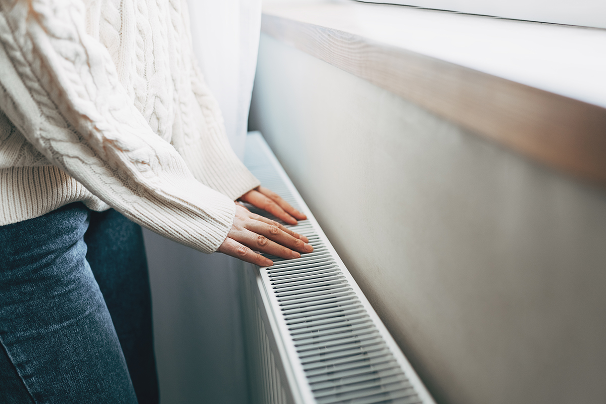 A woman wearing a white knitted jumper and denim jeans has her hands on a radiator next to a window.