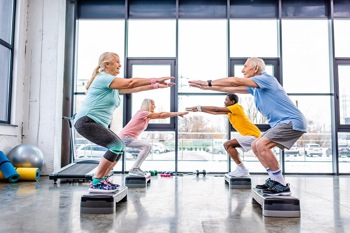 The side view of elderly people doing squats with their arms out in front of them on a step platform in a gym.