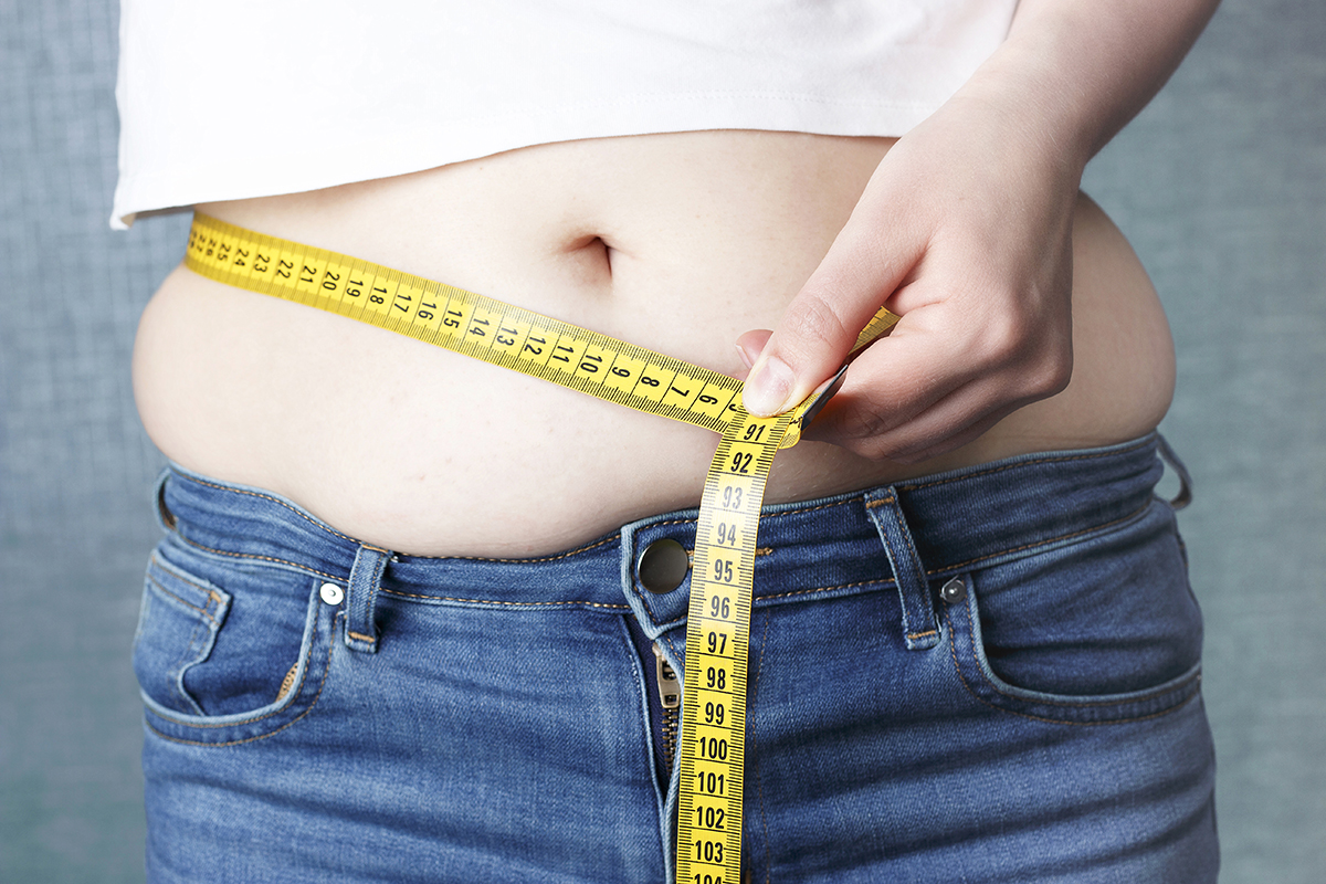 A person holds a yellow measuring tape around their stomach with one hand.