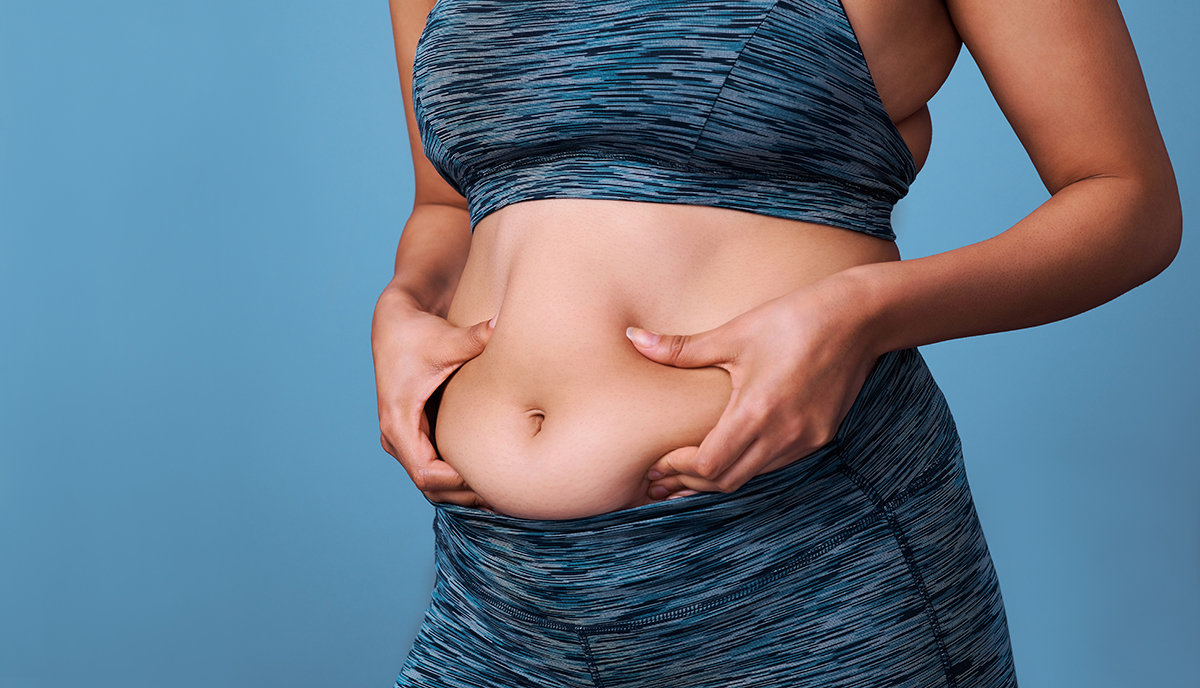 The midsection of a woman wearing an activewear set and pinching the fat on her stomach.