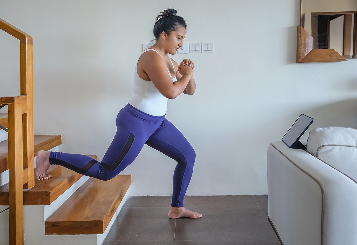 A woman wearing a white vest top and blue leggings does a lunge in her home with her back leg elevated on the second step of the stairs.