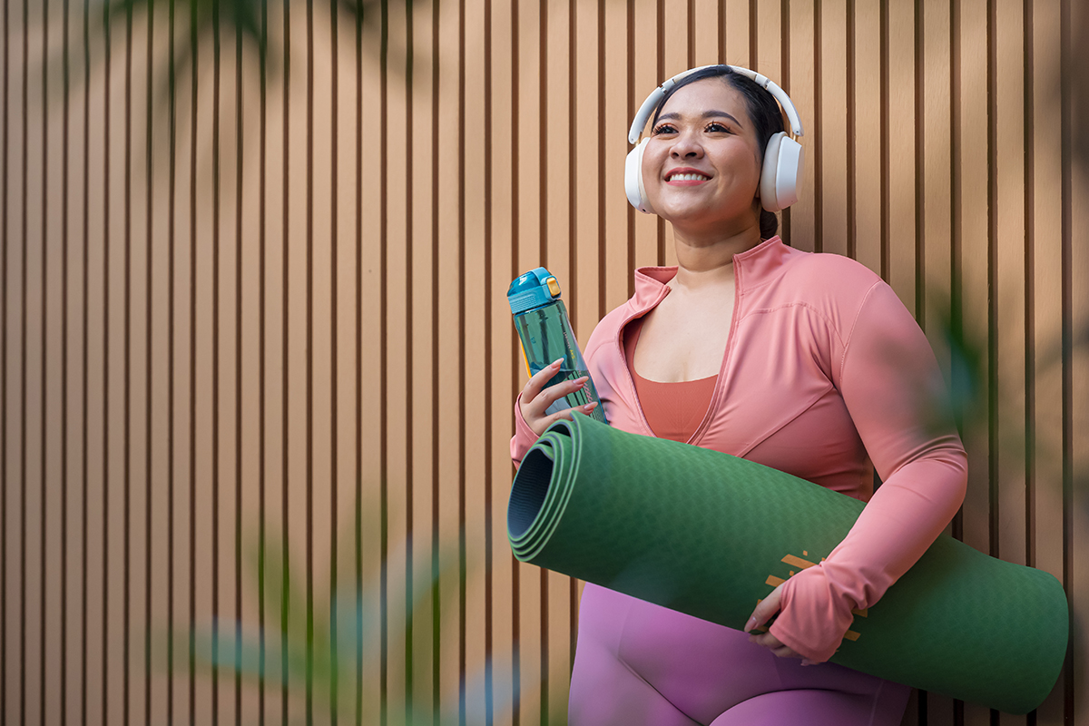 Woman wearing pink and purple workout clothes and headphones holds a green yoga mat and bottle.