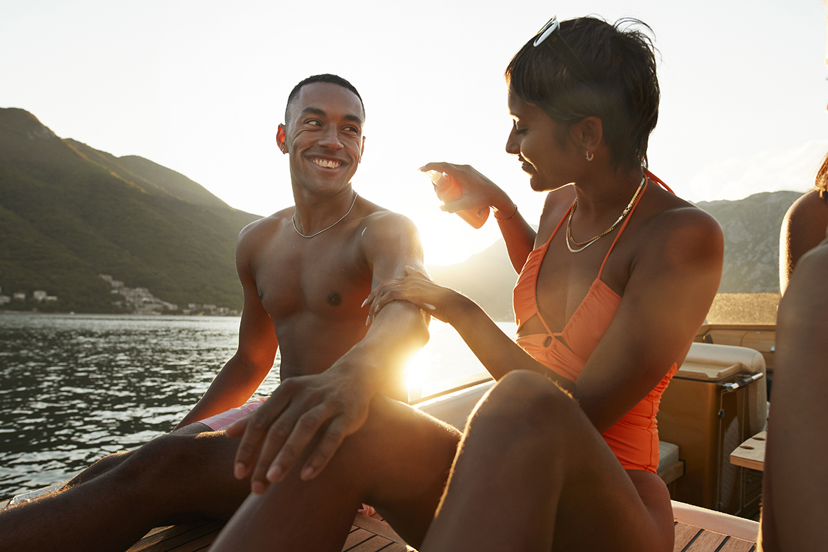 A couple are on a boat at sunset and the woman is applying suncream to the man's arm