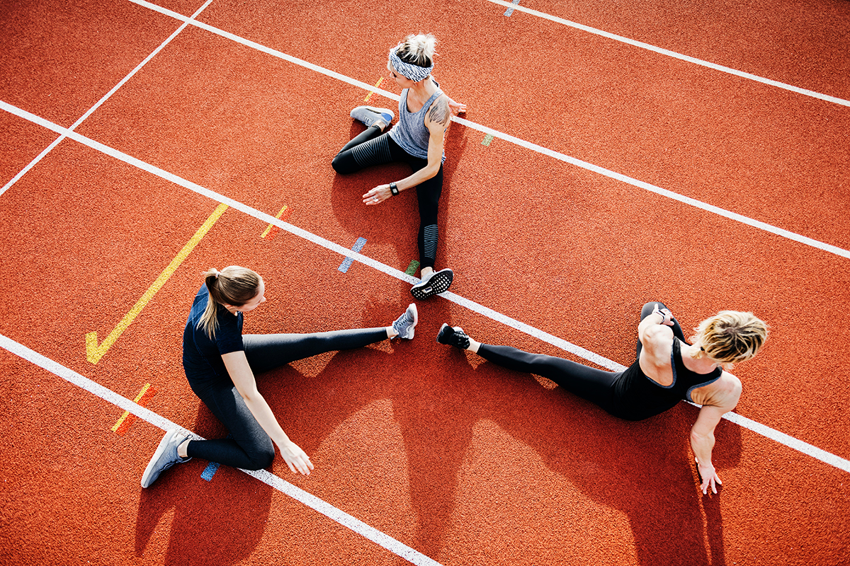 Three women sitting in a single-leg hamstring stretch on a running track