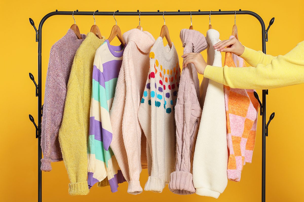 A woman is looking through a selection of jumpers on a closed rail against a yellow background