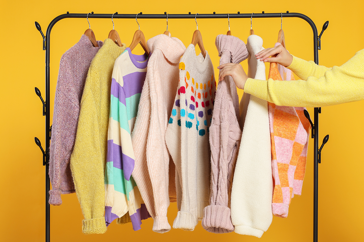 A woman is looking through a selection of jumpers on a closed rail against a yellow background