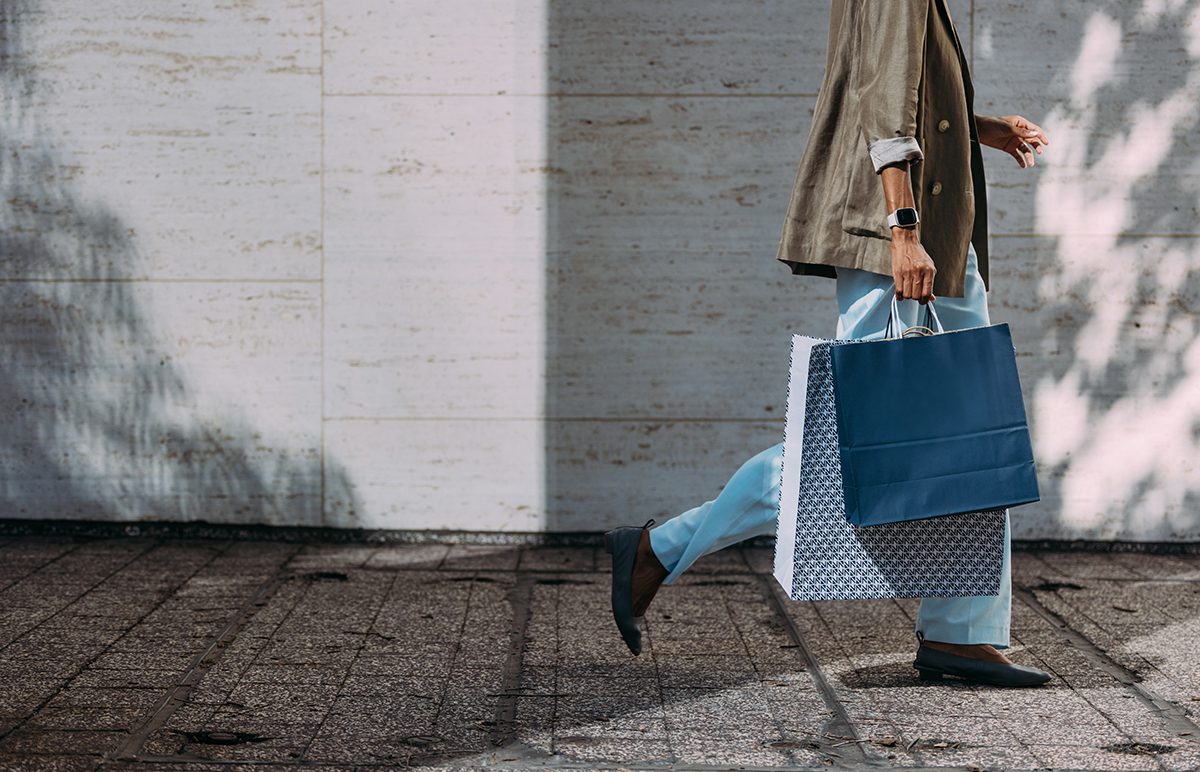 A stylish woman wearing a jacket and jeans walks down the street carrying two large shopping bags