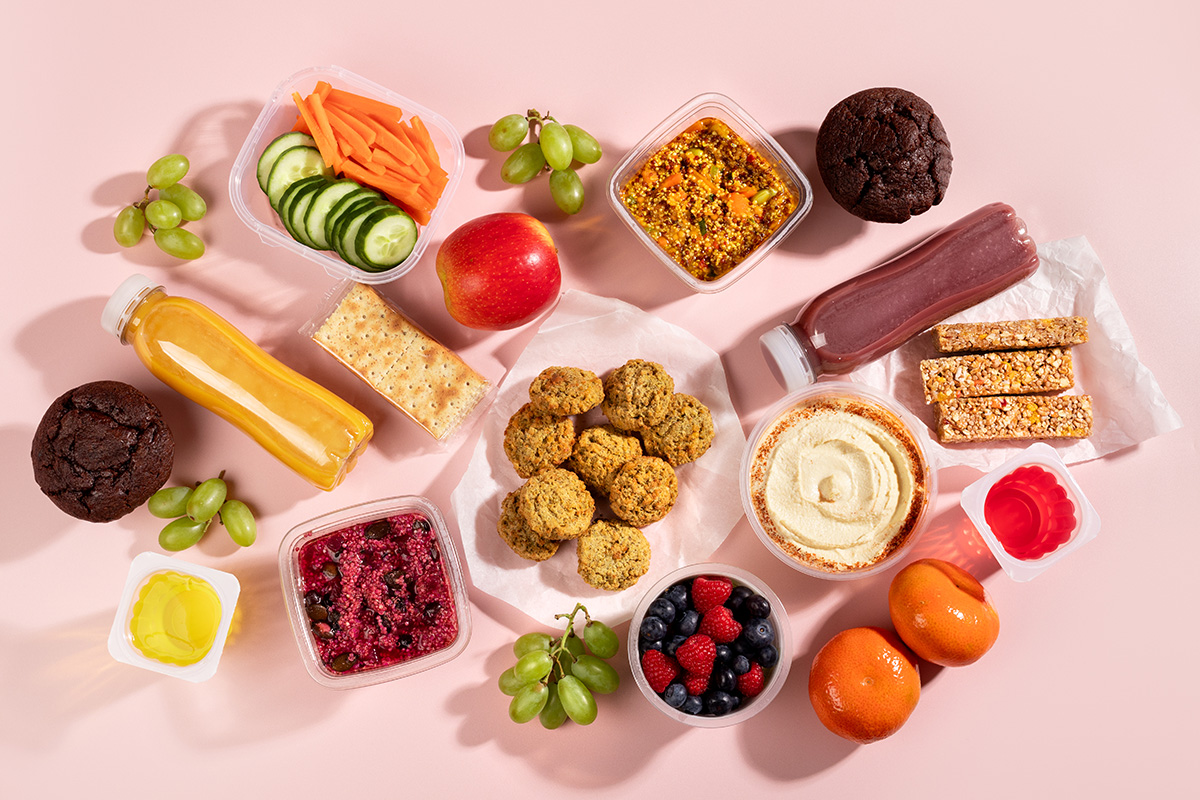 A selection of foods, including fresh fruits and vegetables, smoothies, quinoa salad, falafel, chocolate muffins and houmous, on a pink background