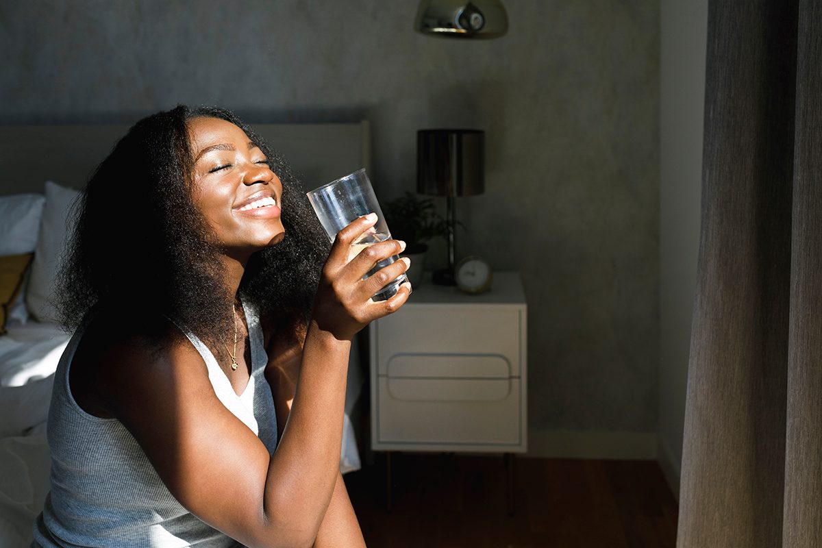 A woman sits in a bedroom holding a glass of water and smiling