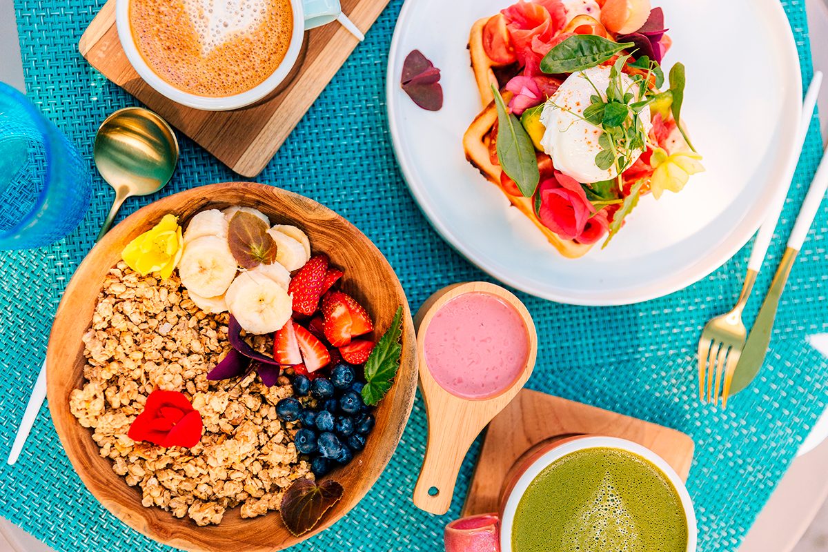 A smoothie bowl, a plate with waffles, smoked salmon and a poached egg on top, a coffee and a matcha sit on a plastic woven placemat