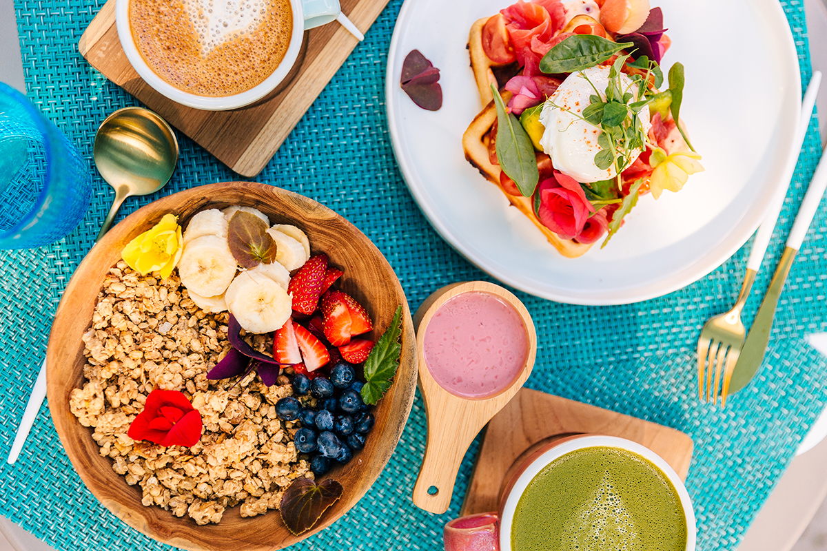 A smoothie bowl, a plate with waffles, smoked salmon and a poached egg on top, a coffee and a matcha sit on a plastic woven placemat