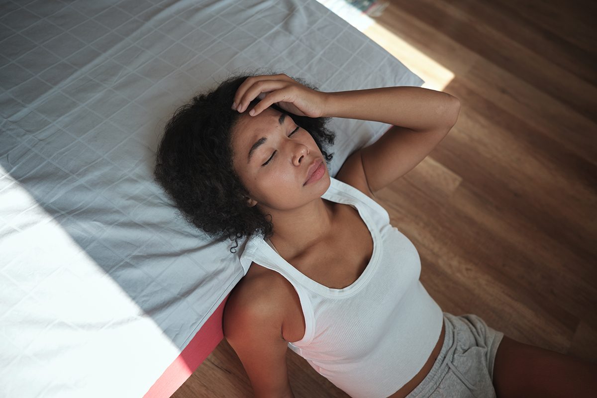 A woman sits on the floor leaning against a bed, touching her forehead