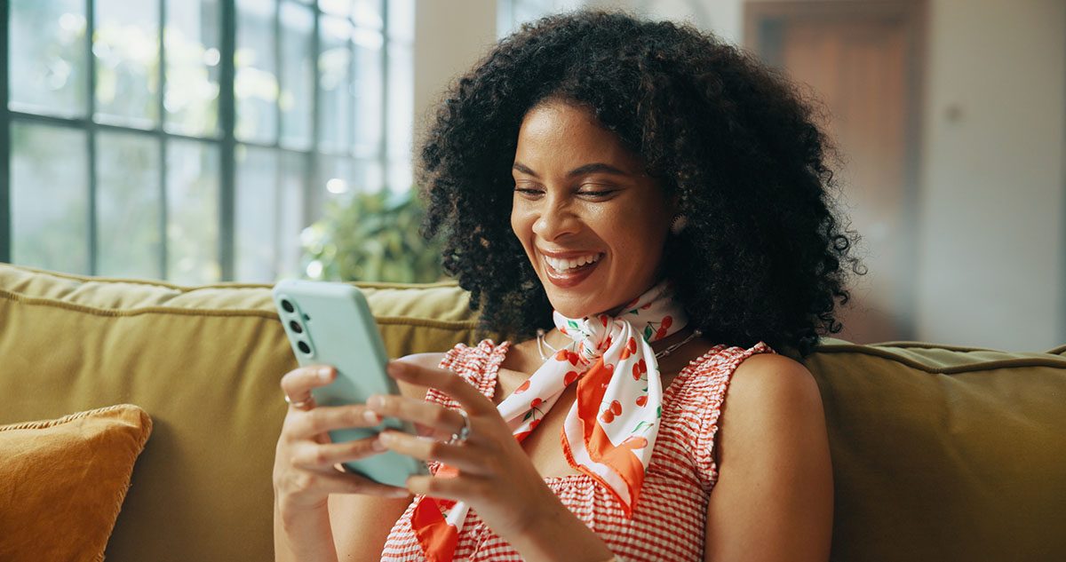 A woman sits on a sofa, looking at her phone and laughing