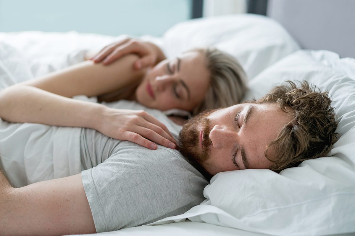 A couple sleep in a bed with white bedding and the woman has her hand on her partner's chest.