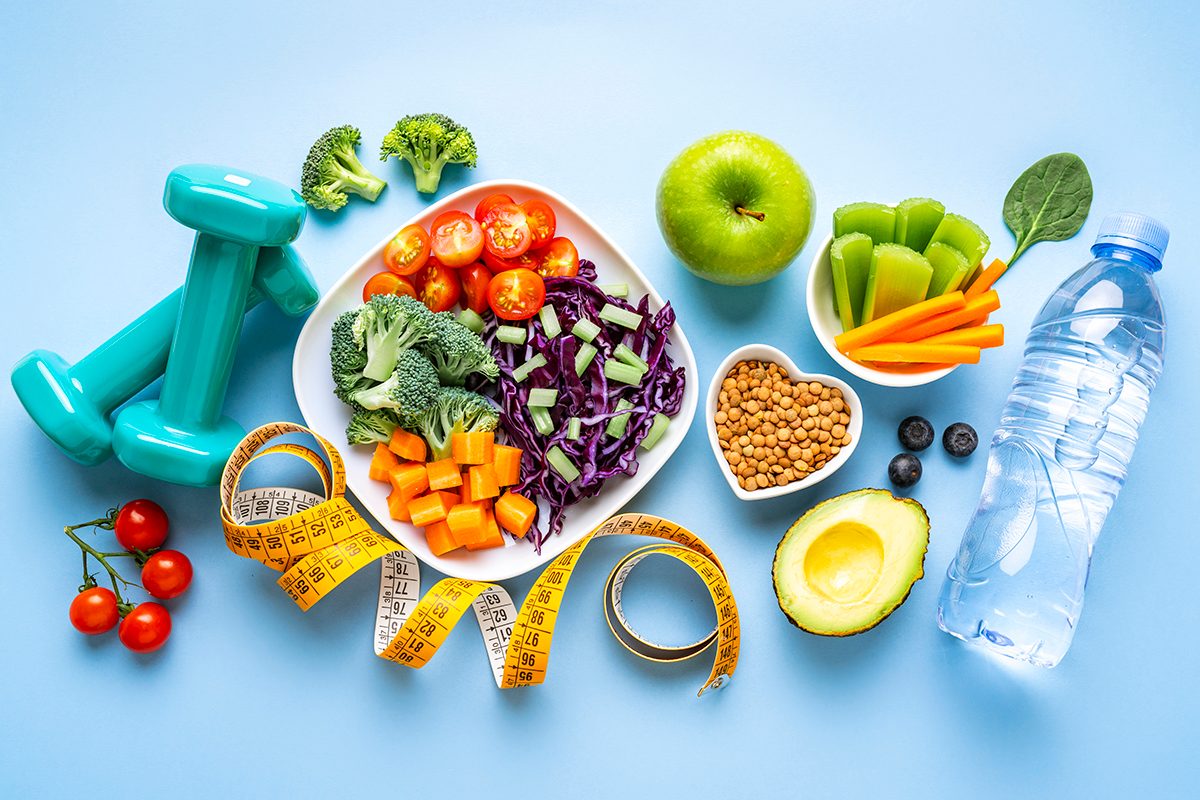 Hand weights, a plate of vegetables, a small heart-shaped bowl of lentils, a bottle of water, and a measuring tape are arranged on a white background.