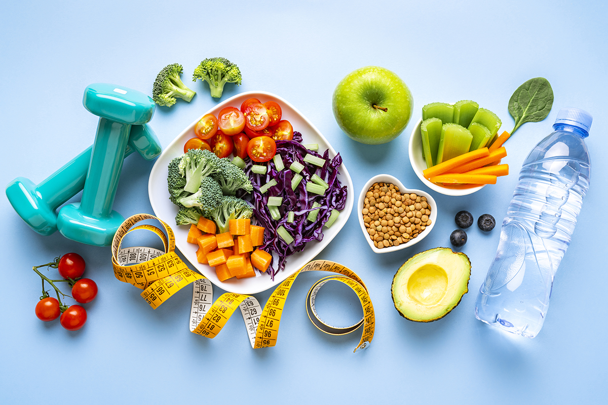 Hand weights, a plate of vegetables, a small heart-shaped bowl of lentils, a bottle of water, and a measuring tape are arranged on a white background.