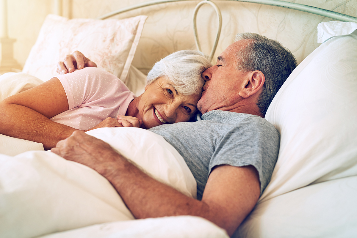 A senior couple cuddling in bed, with the husband kissing his wife's head