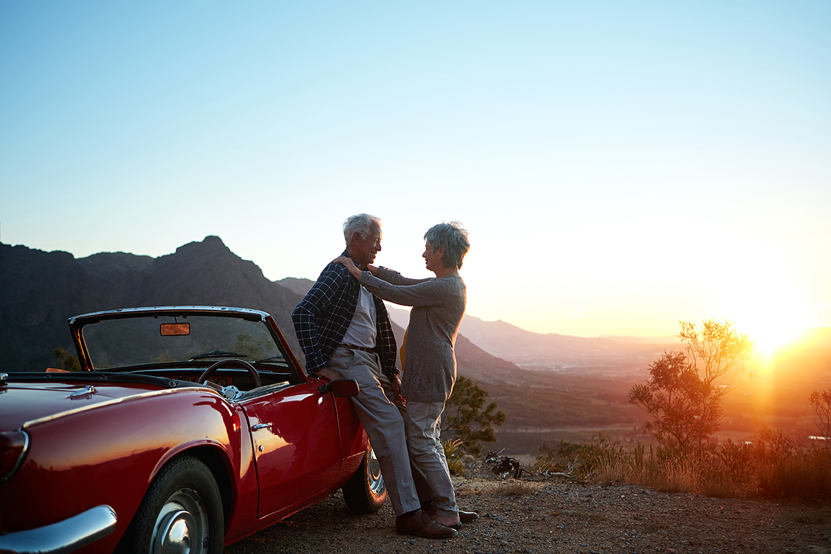 A senior couple stand by a car at sunset, with the wife resting her hands on his shoulders