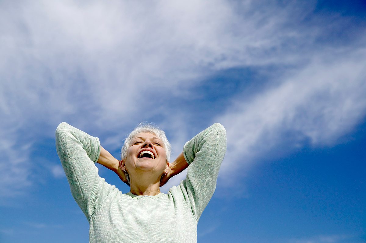 A carefree woman is smiling with her hands behind her head against a blue sky