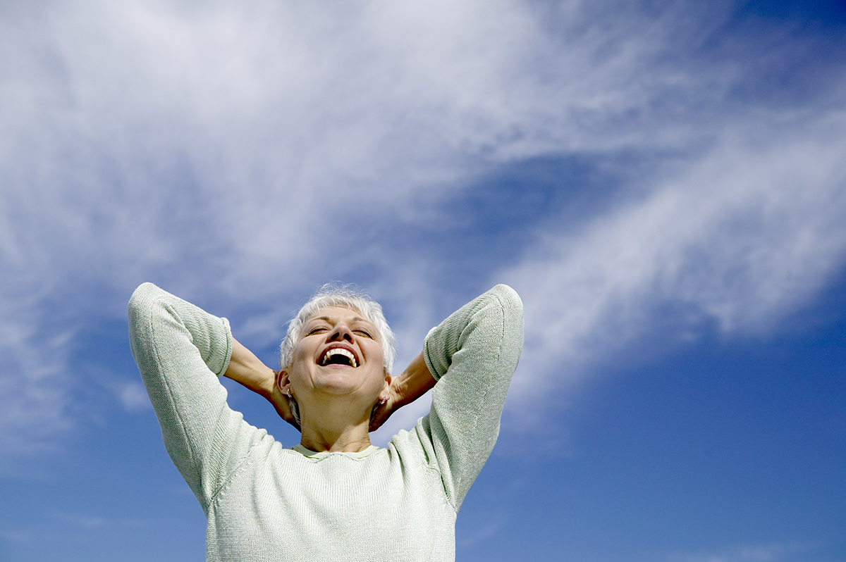 A carefree woman is smiling with her hands behind her head against a blue sky