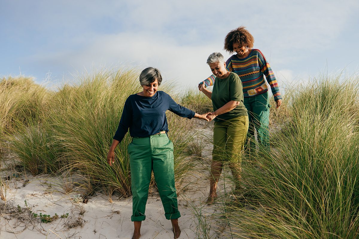 Three middle-aged women are walking across a sand dune holding hands