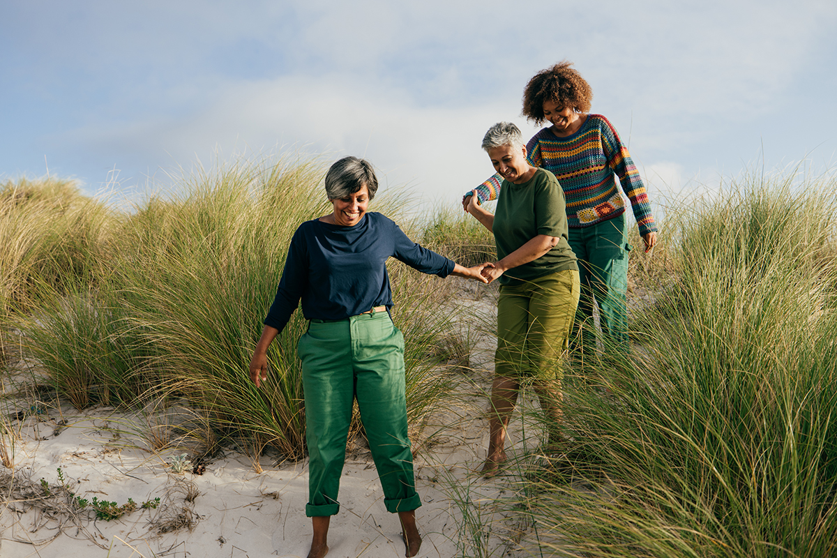 Three middle-aged women are walking across a sand dune holding hands