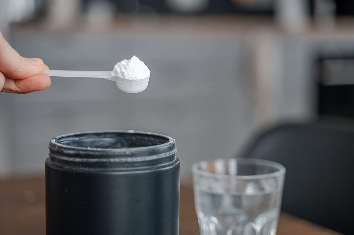A scoop of creatine next to a black tub of creatine and a glass of water